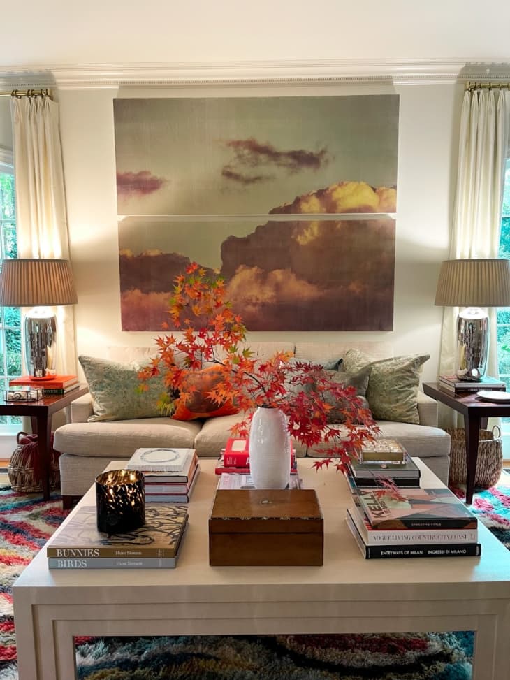 Living room with a beige sofa, colorful rug, large cloud artwork, and a coffee table with books and a vase of autumn leaves.