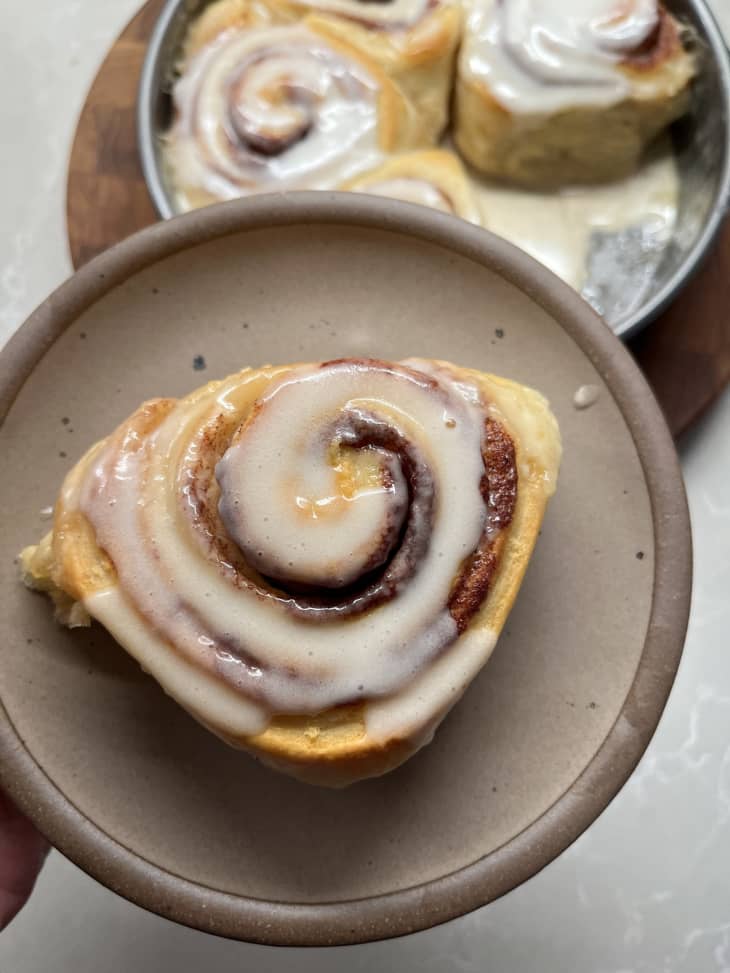 Cinnamon roll with icing on a beige plate, with more rolls in a pan in the background.