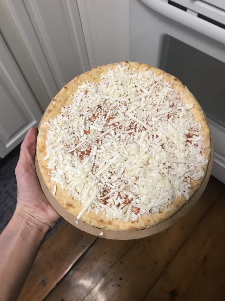 Unbaked cheese pizza held in hand over wooden kitchen floor.