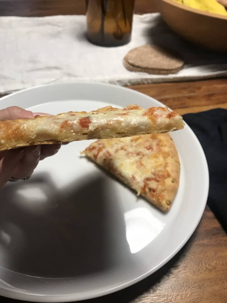 Hand holding a slice of cheese pizza over a white plate with another slice, on a wooden table.