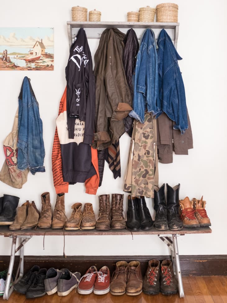 Coat rack with various jackets and coats above a bench lined with boots and shoes, wicker baskets on top shelf.