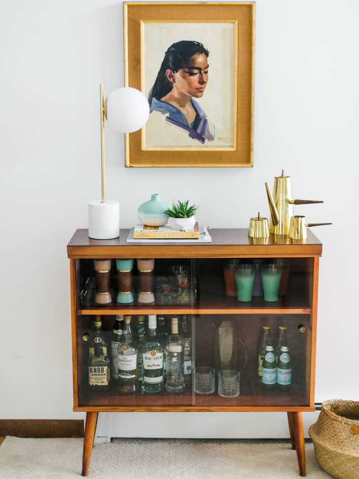 Mid-century bar cabinet with liquor bottles, glassware, and a gold watering can, topped with a lamp and decor, beneath a portrait.