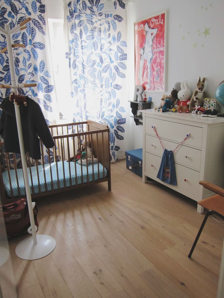 Nursery with wooden crib, blue leaf curtains, white dresser with toys, and a red poster on the wall.
