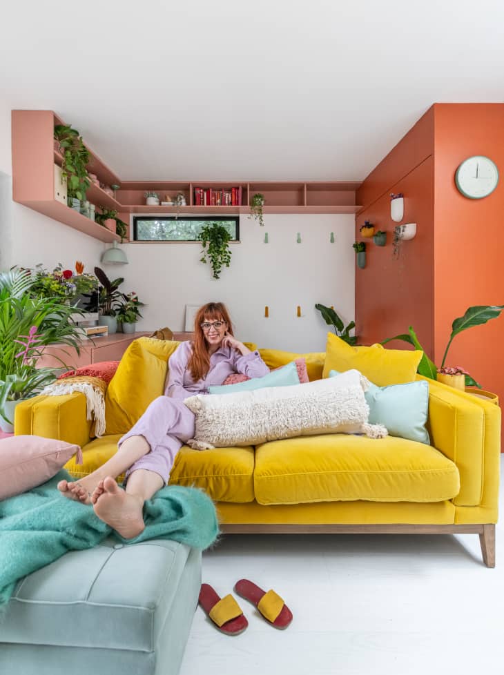 Woman lounging on a yellow sofa with colorful cushions, surrounded by plants and pastel decor in a cozy living room.