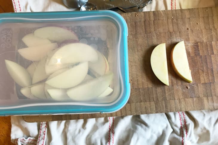 Sliced apples in a blue container on a wooden cutting board with two apple slices beside it.