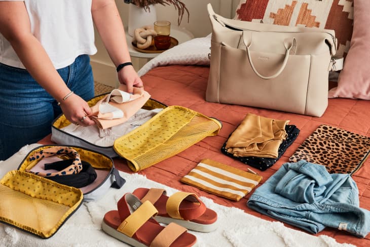 semi close shot of a person packing clothes in a packing cube on a bed with burnt orange sheets and a cream travel tote bag next to it with folded clothes and shoes above it
