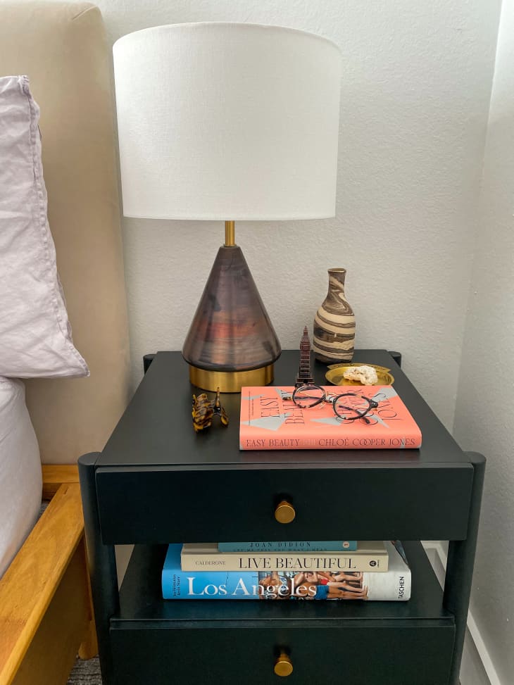 A black nightstand with two drawers and an open shelf in between. There's a lamp with a gold and shiny base beside a book, glasses, a vase, and a gold tray. There's a stack of books on the open shelf. The lamp has a cream-colored base that's smooth in the front.