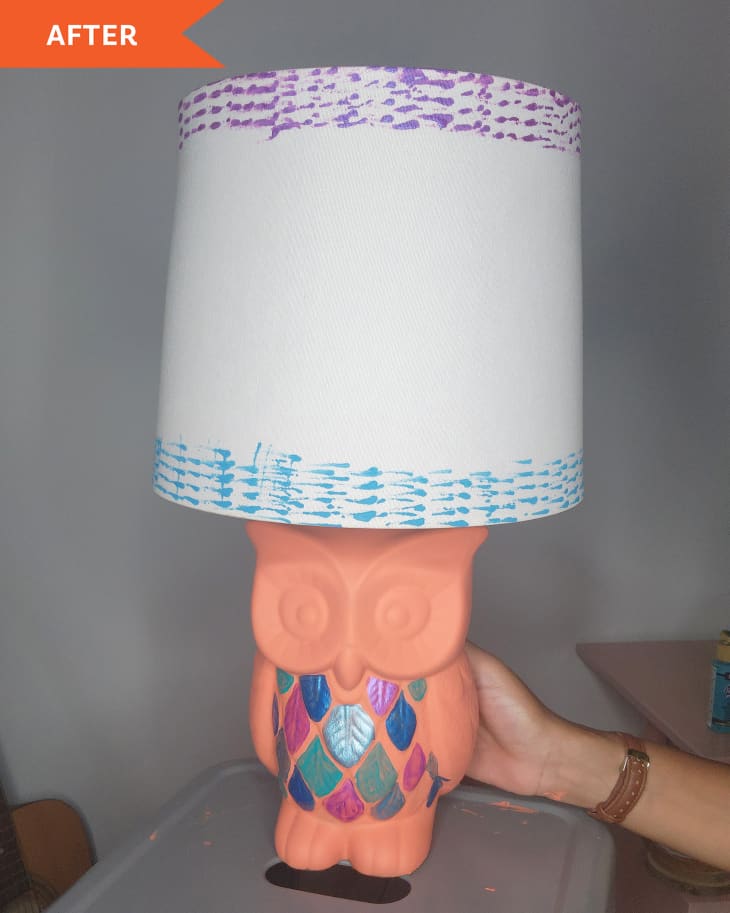 Hand of DIYer holding a terra cotta owl lamp where the feathers have been painted in metallic blues and purples, and the shade has been painted with blue and purple patterns.