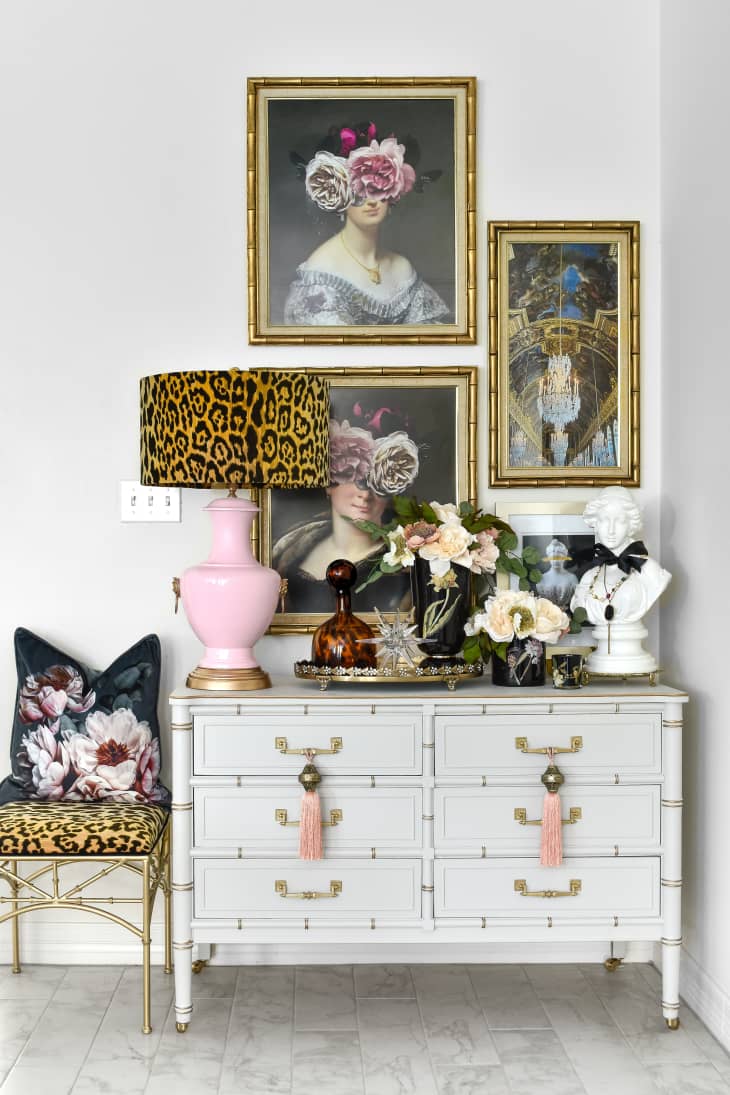 corner of a room with white dresser covered with flowers, a gold tray with a decanter, and a lamp that's been painted with pink paint and has a leopard shade. There is a gold chair with a leopard cushion nearby. Art is hanging on the wall above.