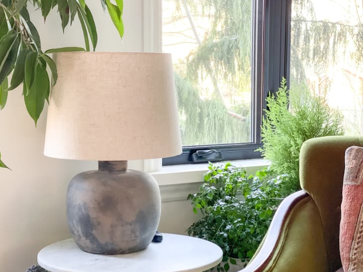 Lamp that has been DIY'ed with a plaster finish sitting on a small round white side table. GReen accent chair can be seen, lush houseplants and window with trees outside