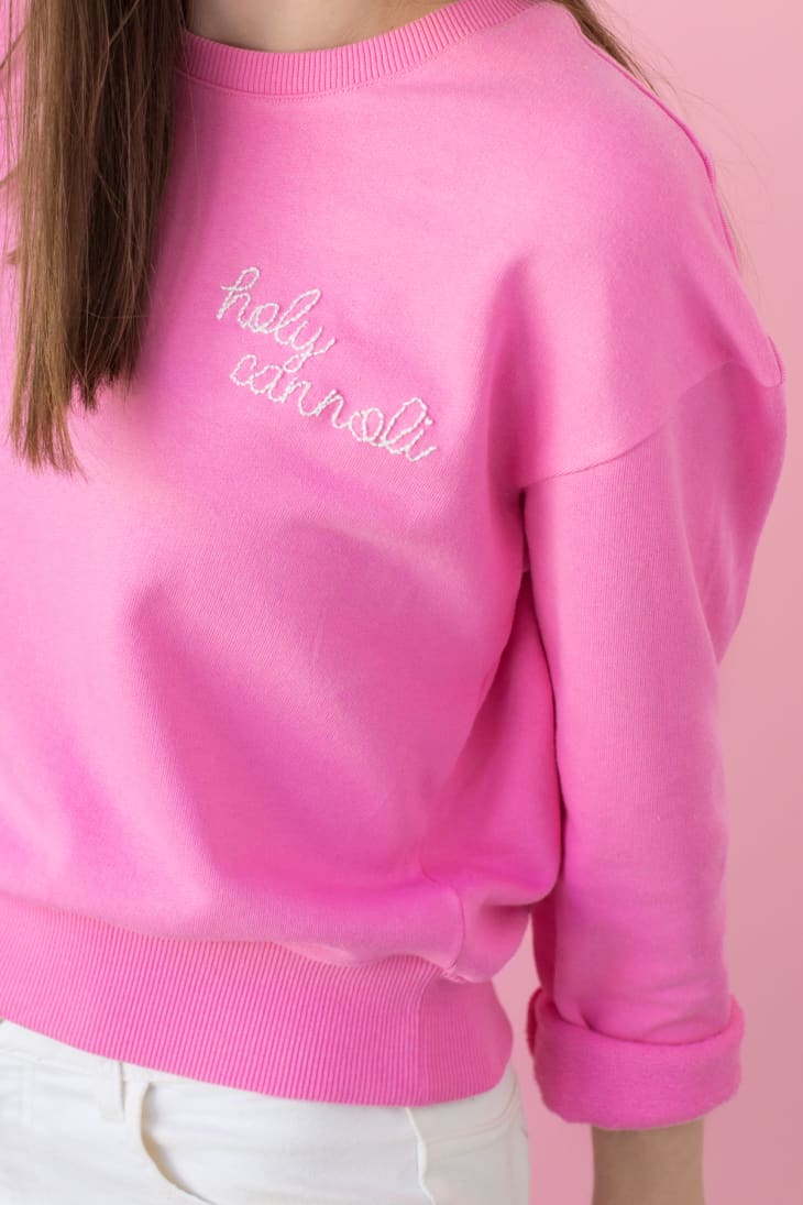 woman wearing a pink sweatshirt embroidered with phrase "holy cannoli"
