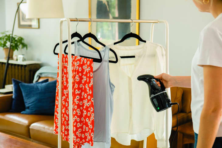 Person steaming clothes on a rack, including a red floral dress, in a cozy living room with a sofa and plant.