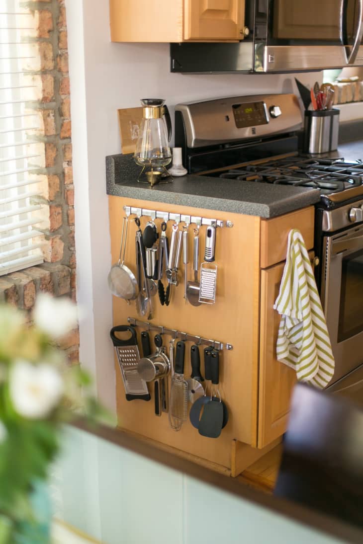 On an exposed side of a wooden cabinet, two rows of rail with hooks are used to hang kitchen tools