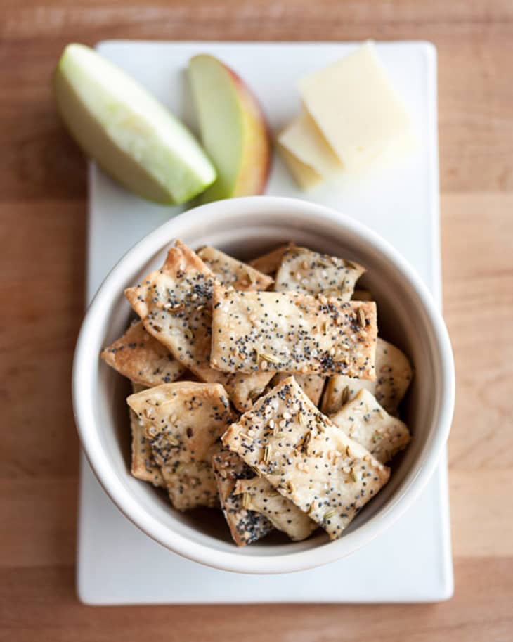 Homemade crackers, topped with fennel seeds, sesame seeds, and poppy seeds, in a bowl, served with apple slices and cheese