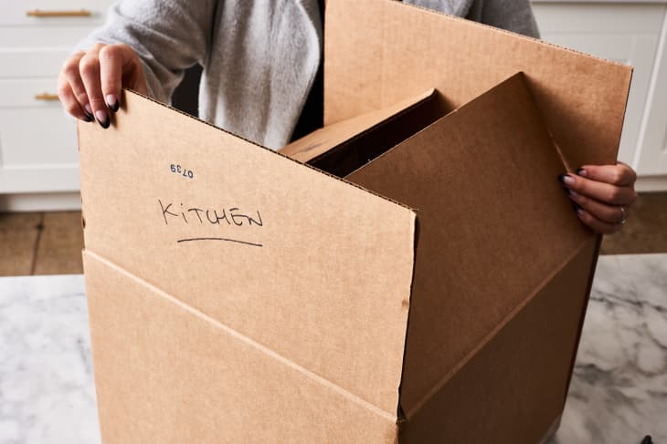 A cardboard box labeled "kitchen" that is being folded to prepare to tape for moving