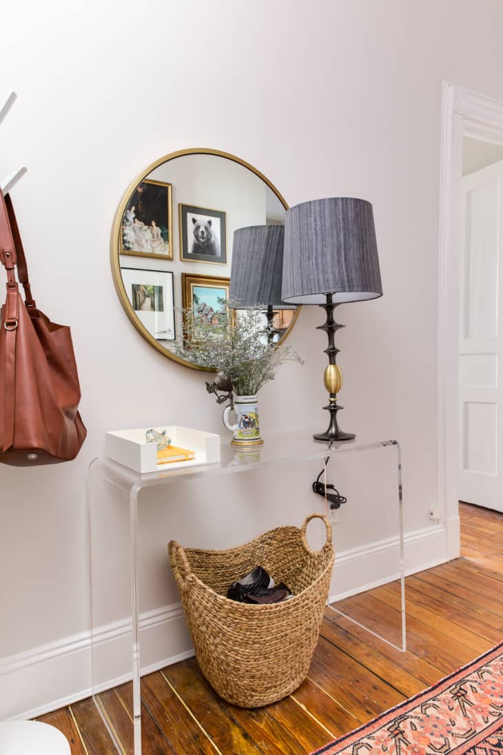 A hallway with a lucite console table with a lamp, plus a basket below and a mirror on the wall