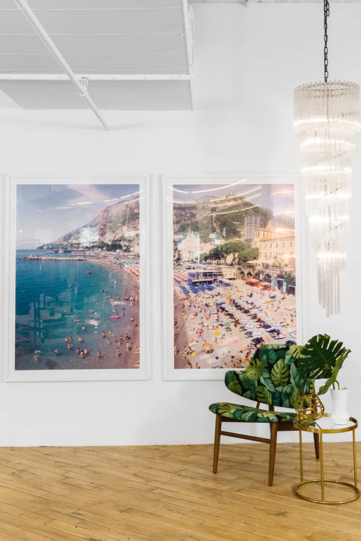 A wall decorated with a diptych of beach photos with a long chandelier and green, monstera-printed chair and brass side table nearby