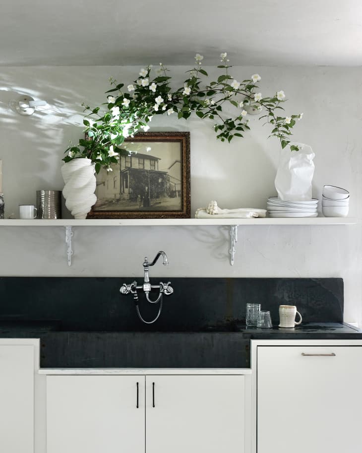 White and black modern kitchen sink/cabinets. White shelf above with white plates, bowls, decorative objets, a framed antique photo, and some white flowers with green leaves arching over the shelf