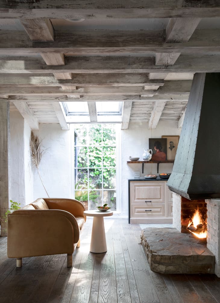 Stone fireplace and sofa, small round table by a kitchen. Exposed whitewashed wood ceiling beams and skylight. Paned window letting in light