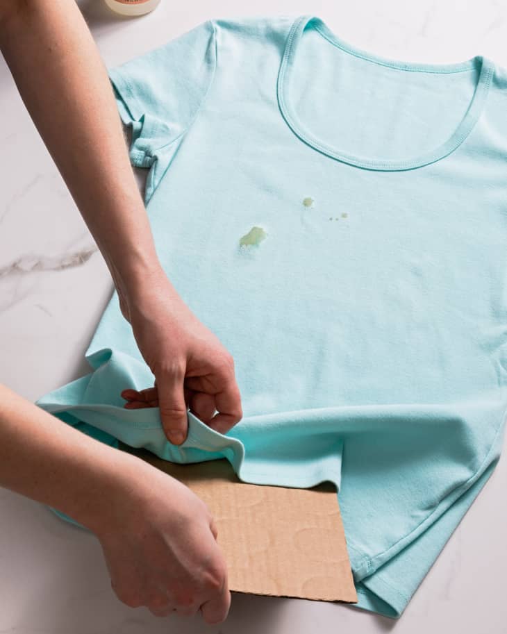 A person placing cardboard between the fabric of a stained t-shirt to prevent the stain from bleeding through
