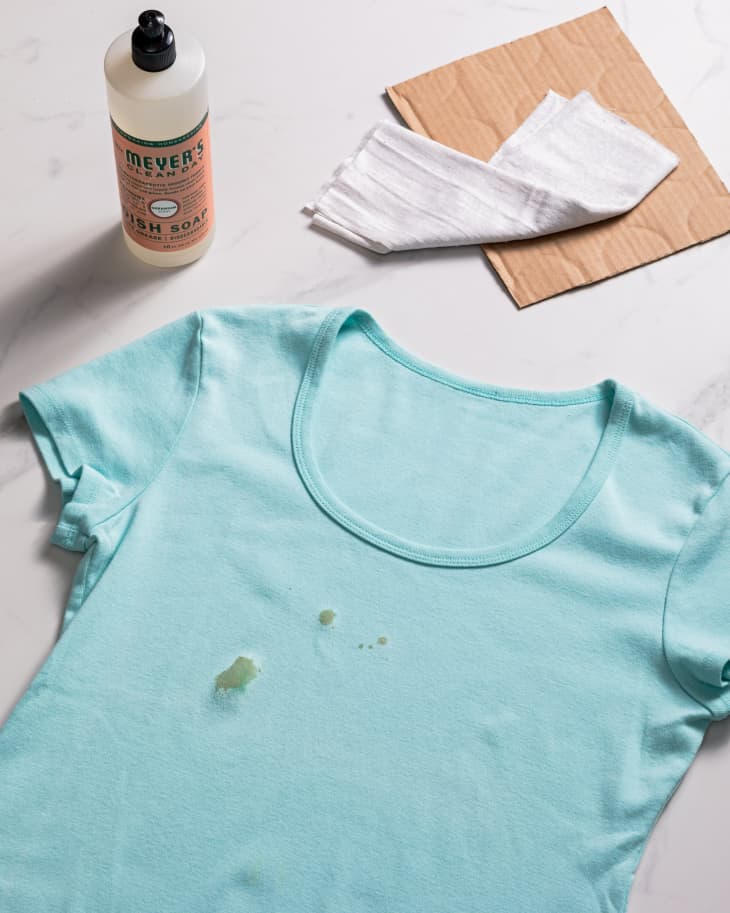 An oil stain on a blue t-shirt, surrounded by cleaning materials like dish soap, cloth, and a piece of cardboard