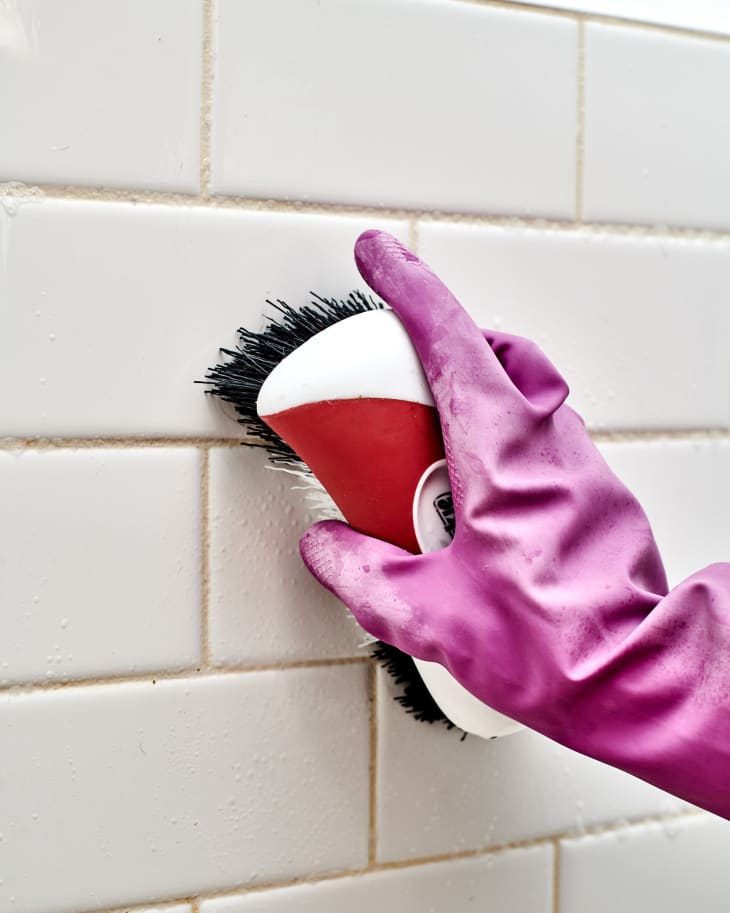 Scrubbing shower walls with a stiff brush wearing dish gloves