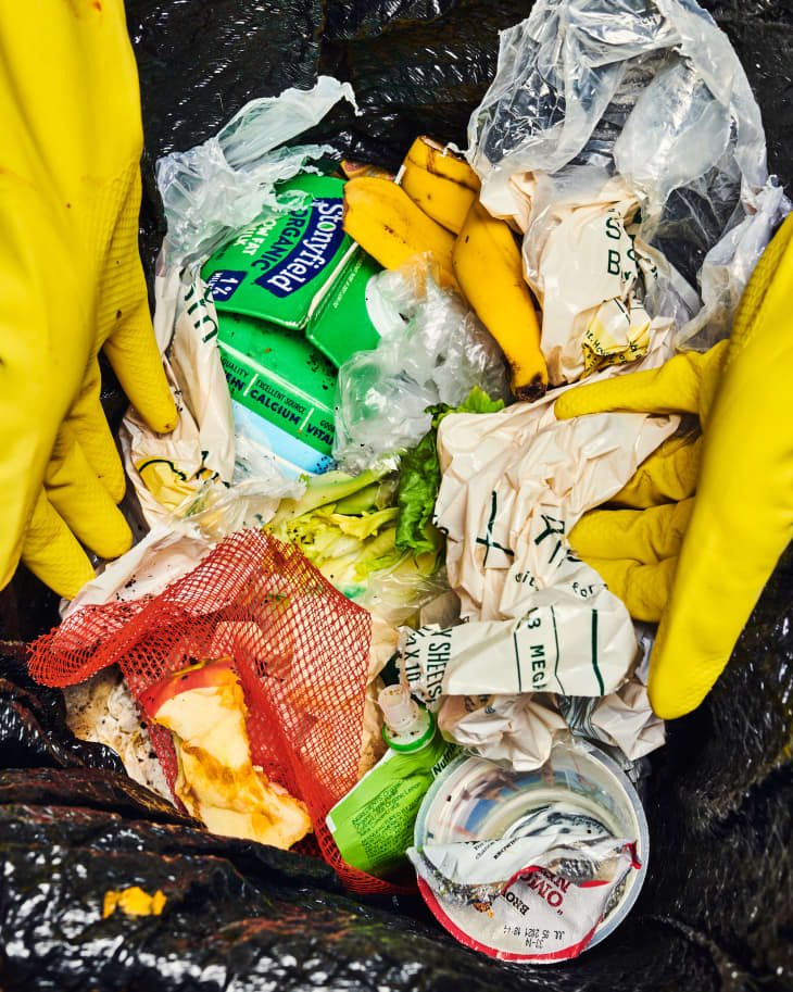 yellow rubber-gloved hand reaching into trash bag where there is an old milk carton, banana peel, yogurt container, and other waste