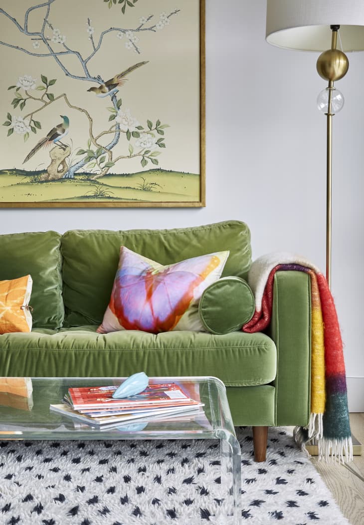 Detail shot of green velvet sofa. Dyed bright-colored throw hanging over one arm. Gold floor lamp with white shade next to sofa. white walls, illustrative bird print on wall behind sofa. Clear lucite coffee table with some magazines. Black and white patterned rug