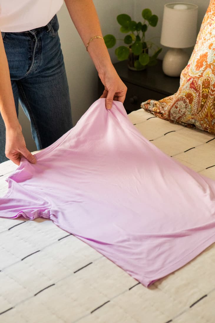 One step showing a woman folding a lavender t-shirt on bed