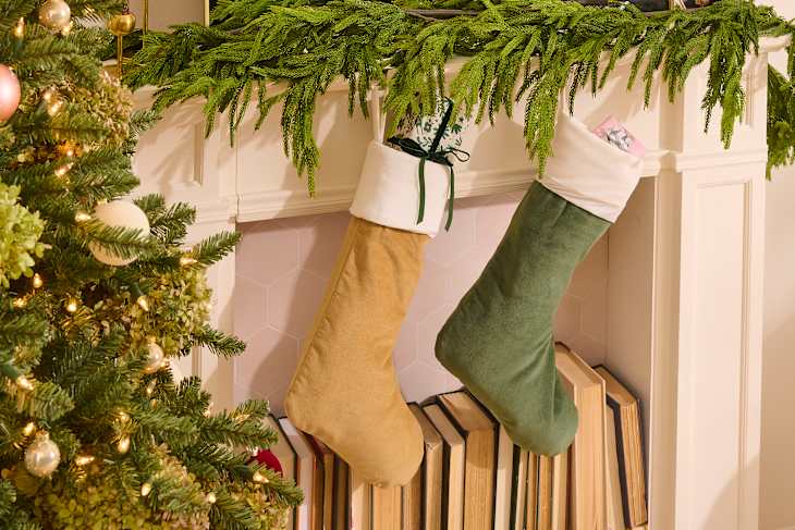 Two festive stockings, one tan and one green, hang from a white mantel adorned with greenery, above stacked books and a Christmas tree.