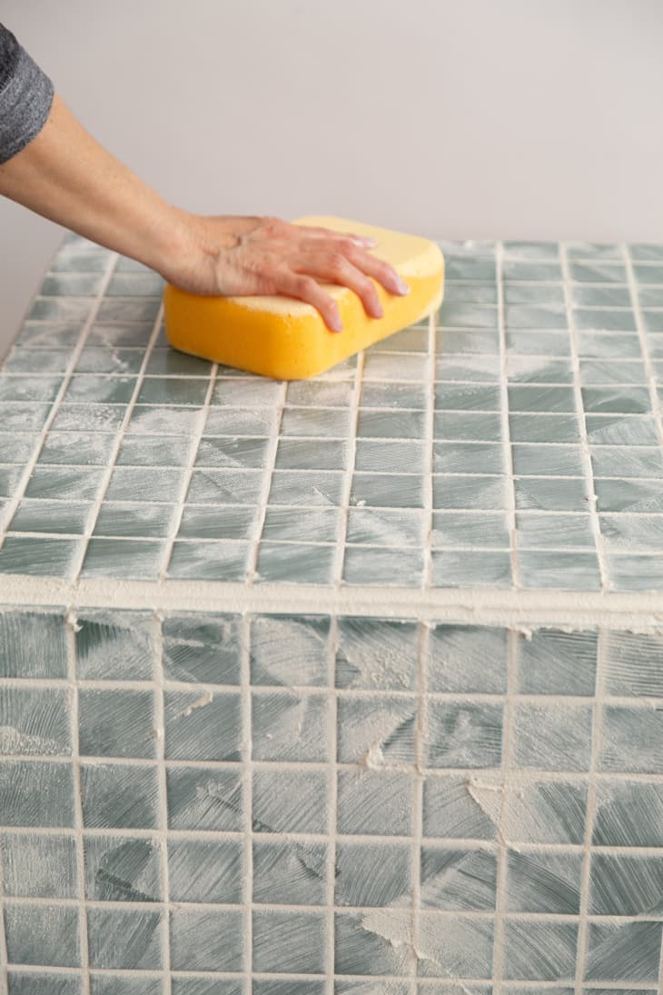Hand holding yellow sponge to wipe off excess grout