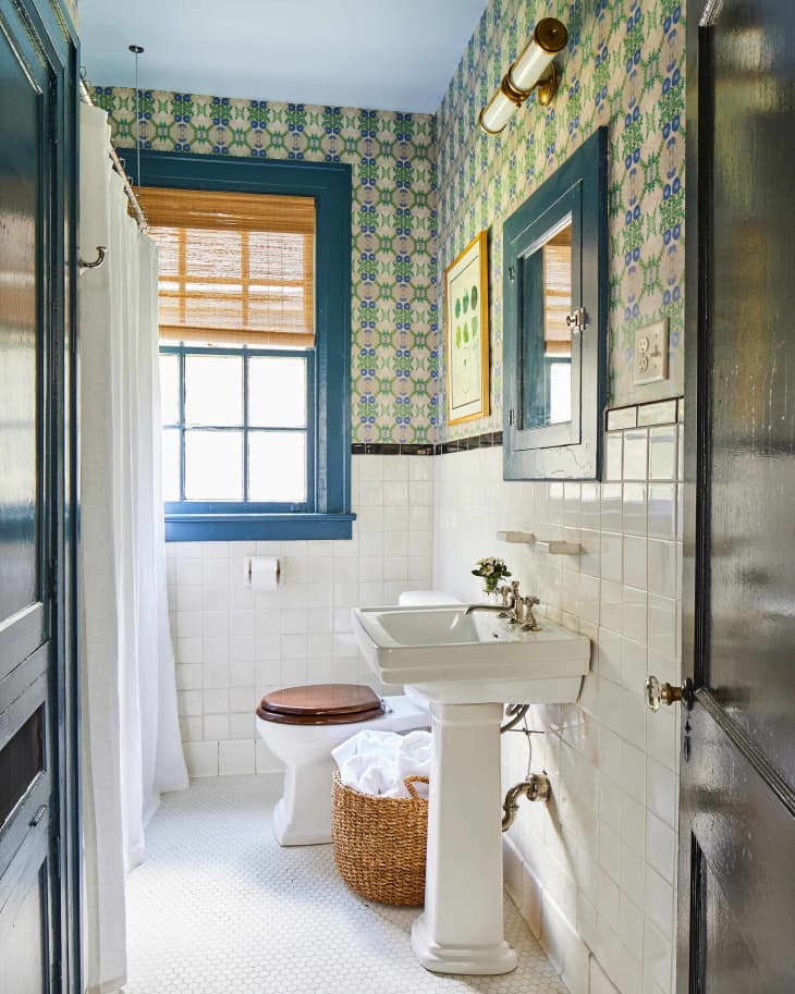 浴室以墙壁为特色，墙壁有一半壁纸，带有彩绘的蓝绿色窗户外壳和医学橱柜框架