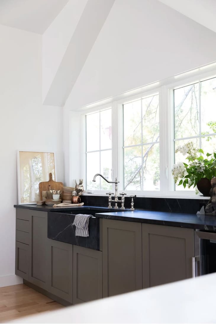 Kitchen with cabinets painted in taupe color, with black countertops and white walls. A black farmhouse sink is in the center of the counter, with a gooseneck faucet.