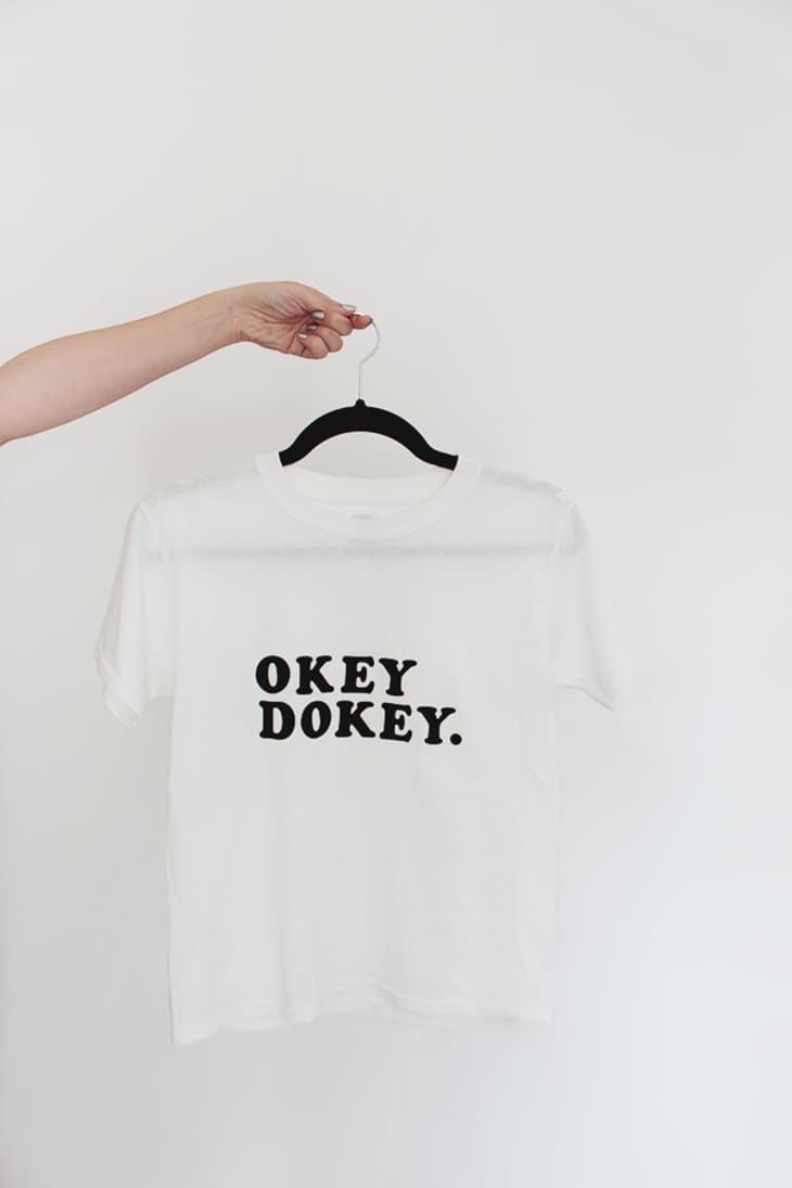 hand holding a white t-shirt with black text saying "okey dokey." on a hanger