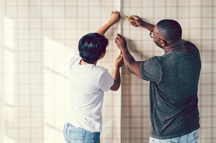 man and woman applying beige plaid-patterned wallpaper to a wall