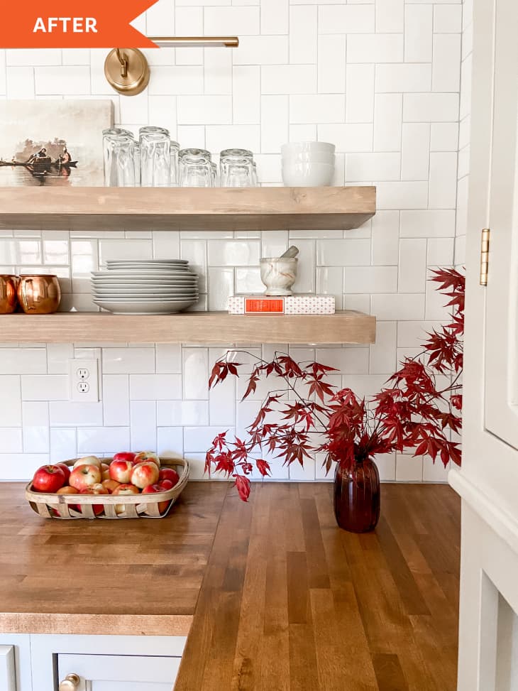 After: corner of kitchen counter with red vase housing red branches and leaves extending out next to floating shelves with plates and glasses on top