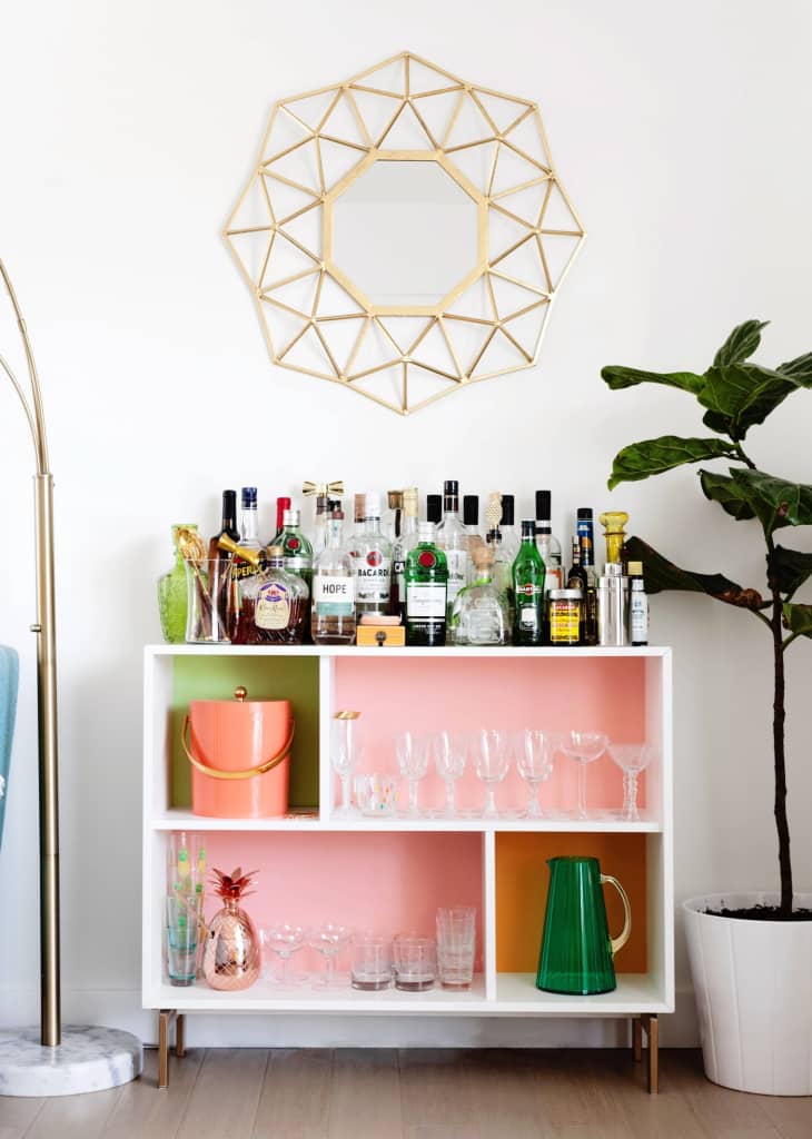 Pink and white bar cart with multiple bottles and glasses next to a plant