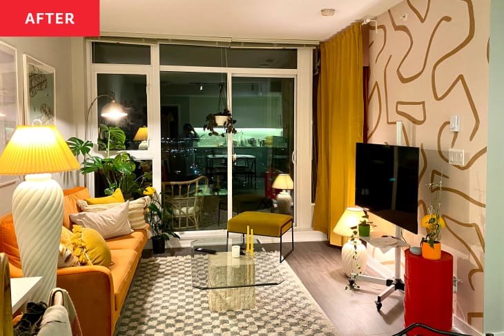 Living room after renovation/makeover. Gold patterned removable wallpaper on wall behind TV, black and white patterned rug, lamps, plants, orange sofa with throw pillows, large windows