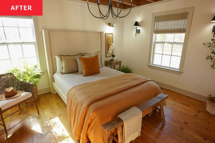 Bedroom after renovation. Hardwood floors, bed with tall fabric headboard, rattan accent chair with white fabric cushion, wood bench at foot of bed, modern chandelier light fixture