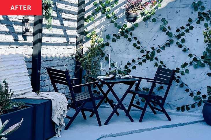 Patio after renovation/makeover. White or cool light gray painted brick and stone wall. Ivy crisscrossing stone wall, black table and chairs, white accent chair with white throw, navy planter box with plants a couple plants in pots