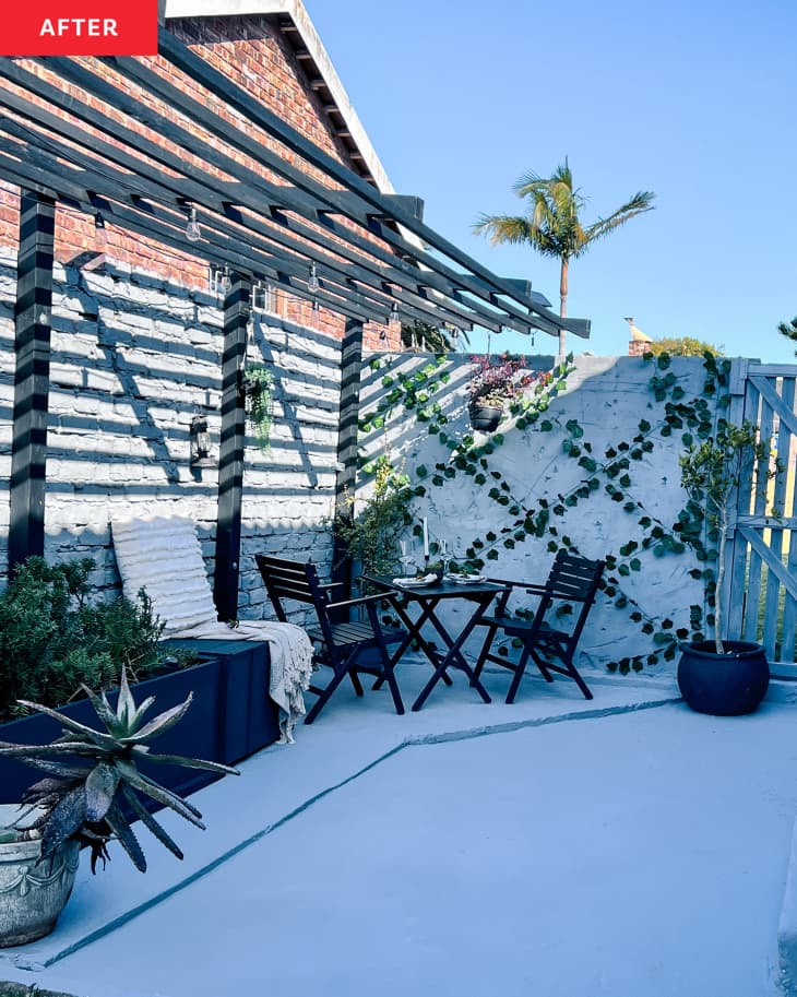 Patio after renovation/makeover. White or cool light gray painted brick and stone wall. Ivy crisscrossing stone wall, black table and chairs, white accent chair with white throw, navy planter box with plants a couple plants in pots
