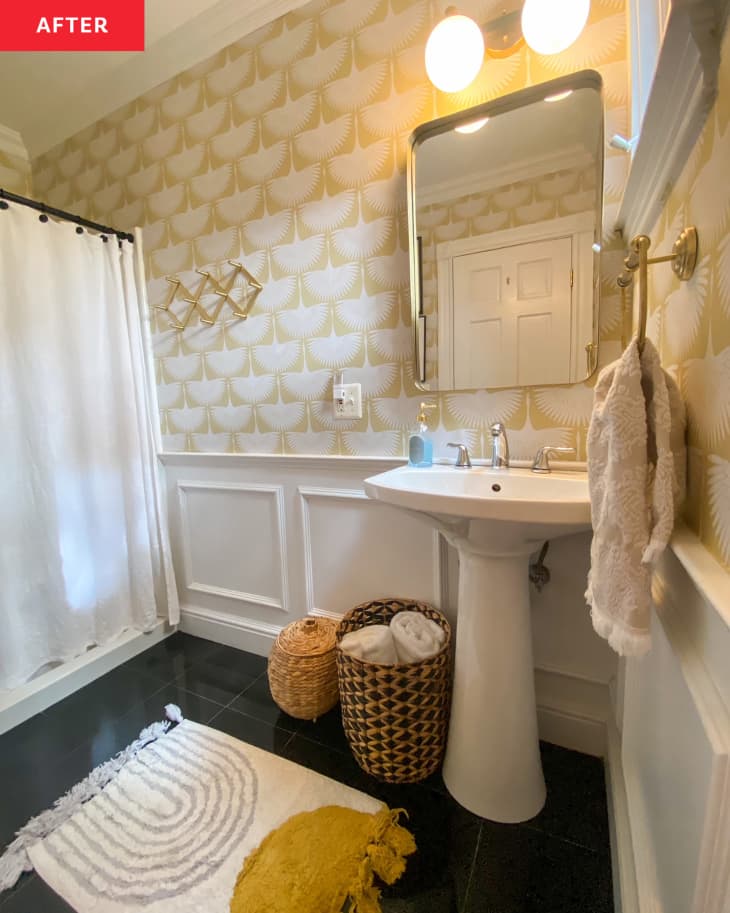 Photo of bathroom after makeover. White and gold patterned wallpaper, gold and black accents, decorative baskets on floor, gold, white and grey shag bath mat