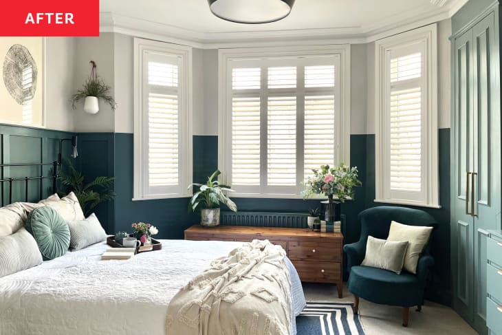 Bedroom after makeover. Bay windows, bottom half of wall around windows painted navy blue, the rest of the room lower half painted green. Upper half of wall around windows is a pale gray. There is a short credenza/dresser under the main window, and a navy blue accent chair with throw pillows. The bed has white, off white linens and there is a breakfast tray with flowers. Tall lighter blue/green cabinets on the right side of the room