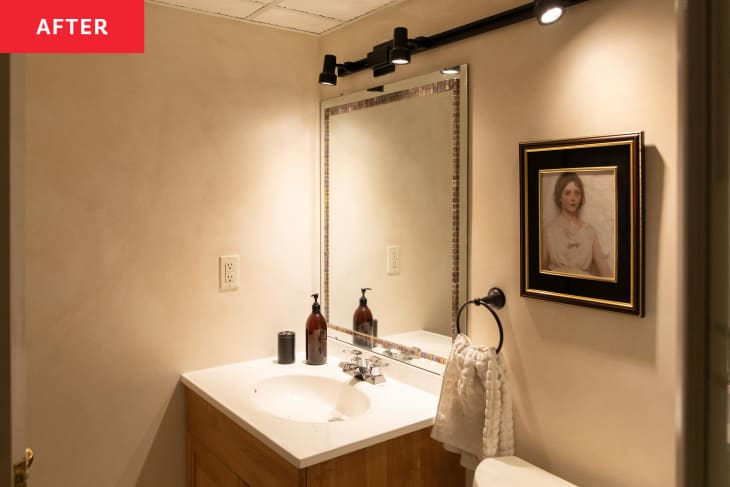 Neutral colored bathroom with vintage painting on the wall and track lighting above mirror.