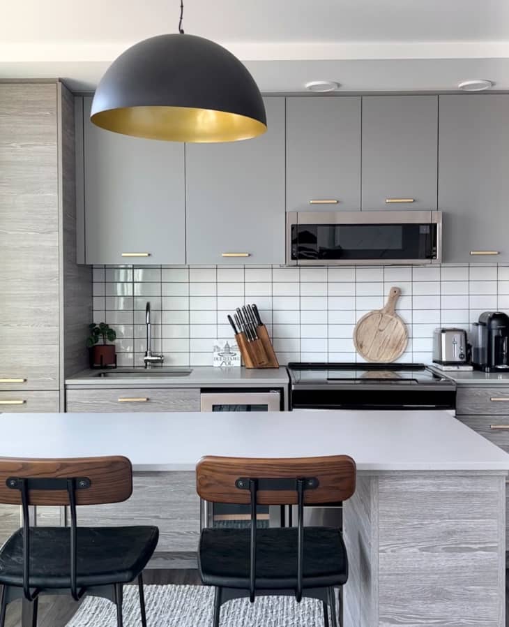 Kitchen with gray cabinets, white tile backsplash, island with 2 wood and black barstools, white ceiling, black pendant lamp over island