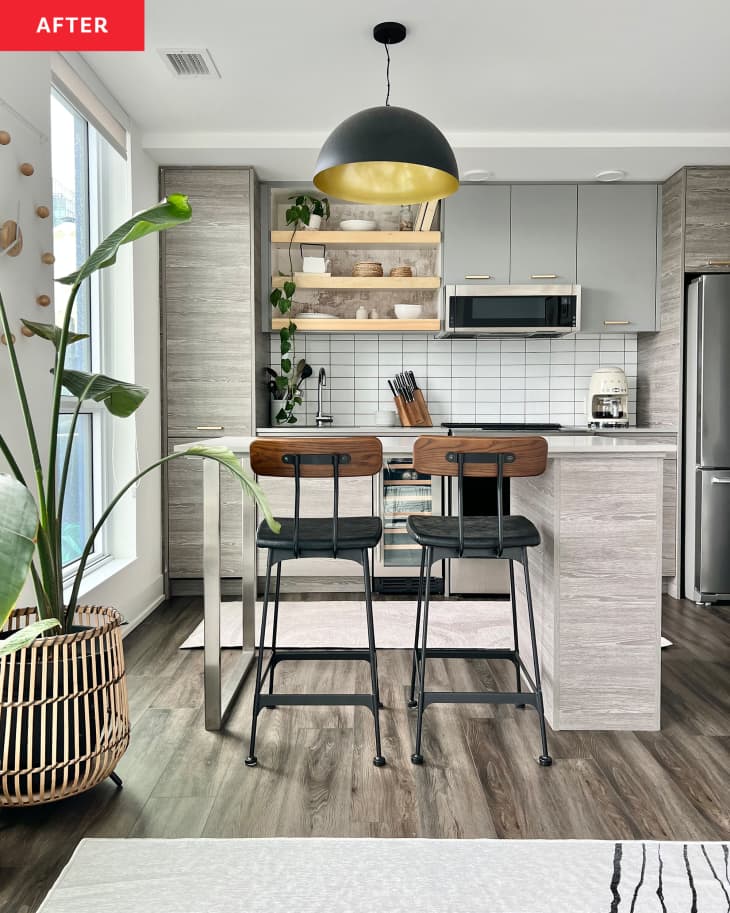 after: Kitchen with gray cabinets, set of open light wood shelves above sink, white tile backsplash, island with 2 wood and black barstools, large tree in basket on floor, white walls, black pendant lamp over island