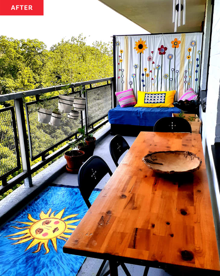 Balcony after makeover/renovation with long wood table with black chairs, blue mat with sunshine design, blue small sofa/daybed with pink and yellow pillows in front of partition covered with handmade wall hanging of flowers made from fabric scraps