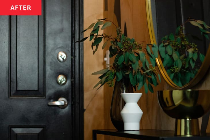 Entryway after makeover: detail of black console table with gold framed round mirror above. Plant in white vase, gold bowl on table. Black door to the left