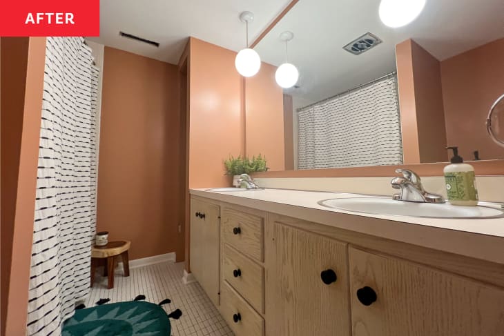 Bathroom after renovation: terra cotta accent walls, white shower curtain with black stripes, black and gray bathmat with star pattern, white small rectangular floor tiles, wood cabinets with black pulls, double sink with hanging globe pendant lights