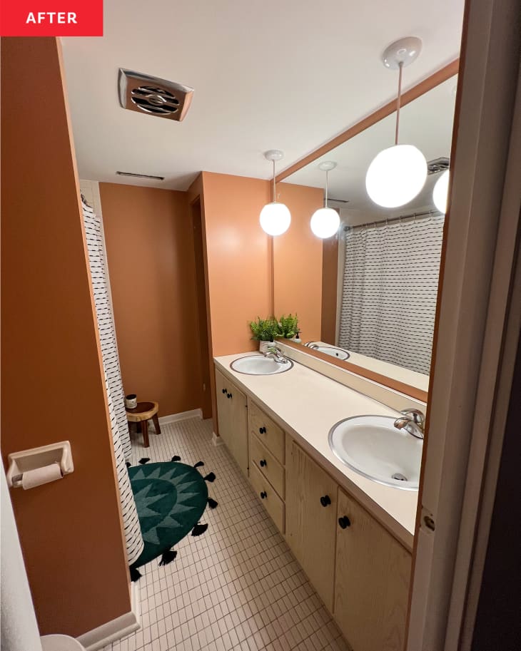 Bathroom after renovation: terra cotta accent walls, white shower curtain with black stripes, black and gray bathmat with star pattern, white small rectangular floor tiles, wood cabinets with black pulls, double sink with hanging globe pendant lights